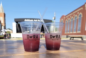 Two DORA cups filled with beverages from a local vendor sit atop a picnic table at the Schine's Art Park. The Schine's Art Park stage sits in the background.